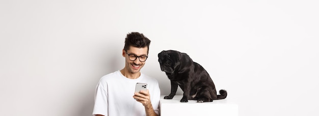 笑顔の若い男がスマートフォンを使用し、犬のパグの所有者の近くに座って、携帯電話で写真をチェックしています。