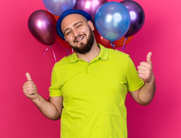Улыбающийся молодой человек в синей партийной шляпе, стоящий перед воздушными шарами, показывает палец вверх, изолированный на розовой стене