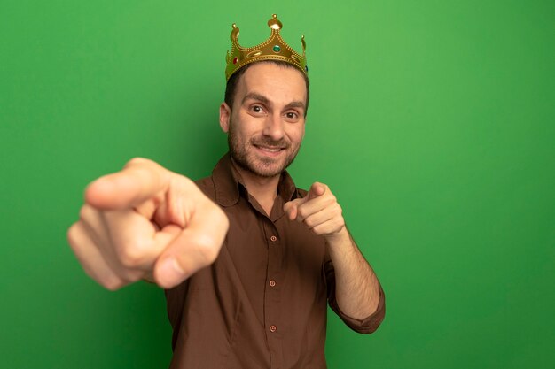 Smiling young man wearing crown looking at front doing you gesture isolated on green wall