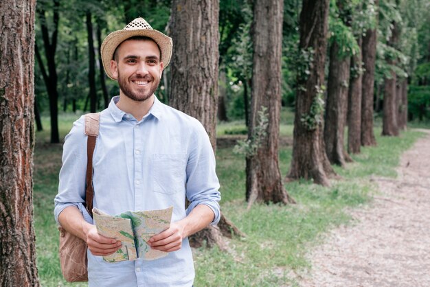 smiling young man wearing hat holding map in park