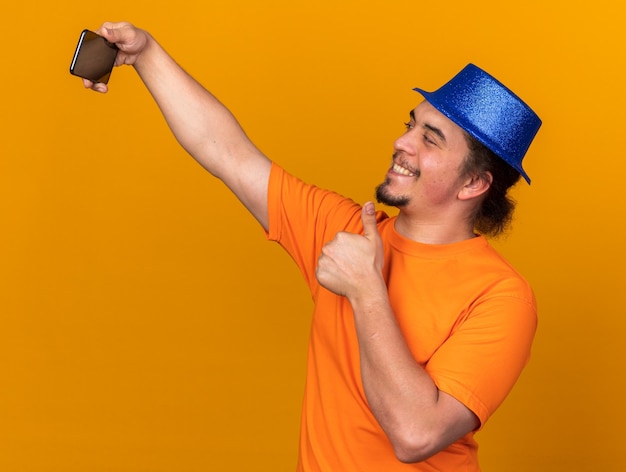 Улыбающийся молодой человек в партийной шляпе делает селфи, показывая большой палец вверх на оранжевой стене