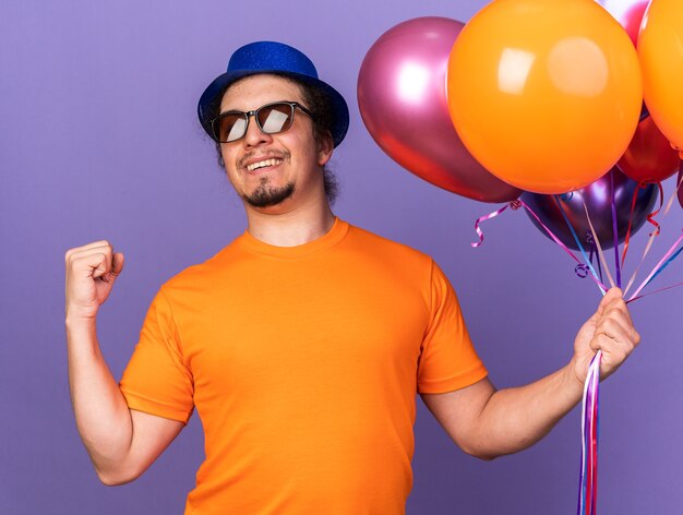 Smiling young man wearing party hat with glasses holding balloons showing yes gesture isolated on purple wall