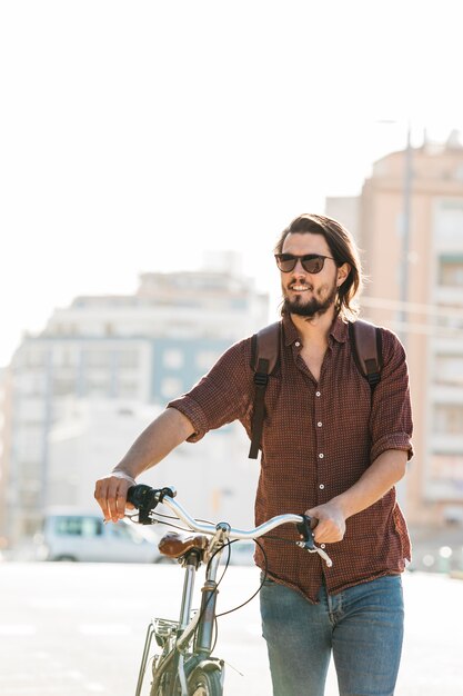 Усмехаясь солнечные очки молодого человека нося гуляя с велосипедом на дороге во второй половине дня