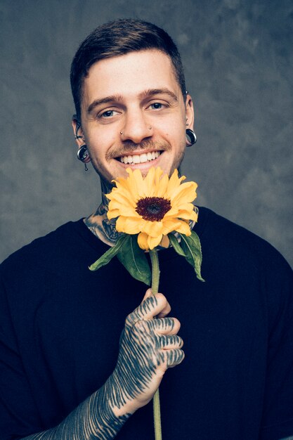 Free photo smiling young man with pierced ears and nose holding sunflower in hand looking to camera