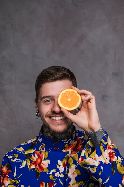 A smiling young man with piercing in the ears and nose holding slice of orange in front of his eyes against grey background