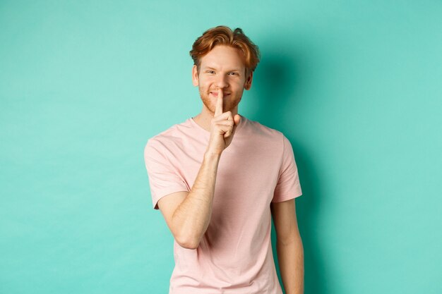 Smiling young man with red hair and beard sharing a secret, showing taboo gesture and grinning, shushing you to be quiet, standing over turquoise background.