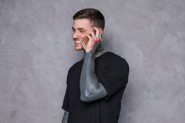 Smiling young man with tattoo on his hand talking on mobile phone against grey wall