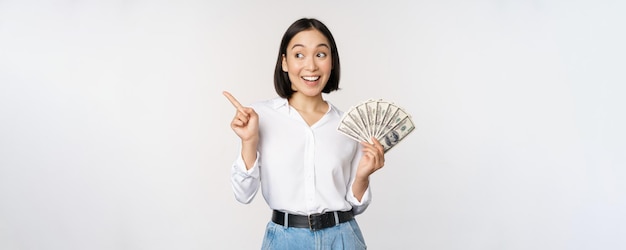 Smiling young modern asian woman pointing at banner advertisement holding cash money dollars standin