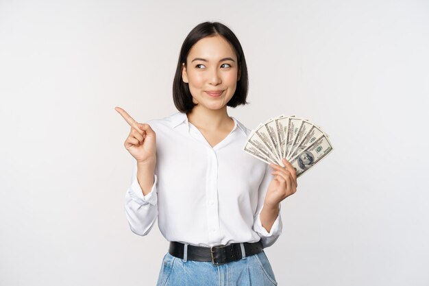 Smiling young modern asian woman pointing at banner advertisement holding cash money dollars standing over white background