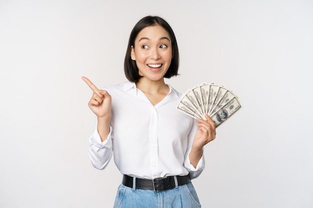 Free photo smiling young modern asian woman pointing at banner advertisement holding cash money dollars standing over white background