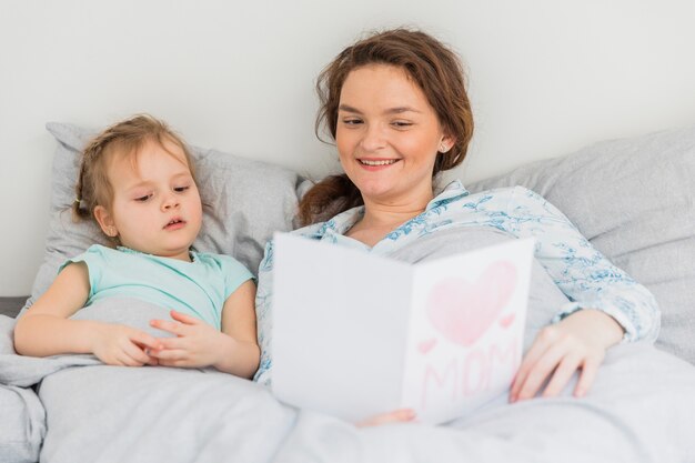 Улыбающаяся молодая мать читает открытку возле своей дочери, лежа на кровати