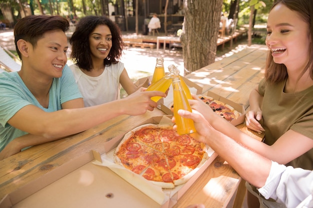 Smiling young multiethnic friends students outdoors drinking juice eating pizza.