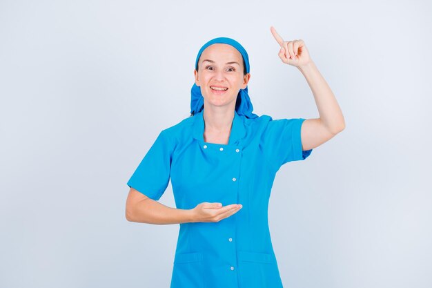 Smiling young nurse is pointing up with forefinger on white background