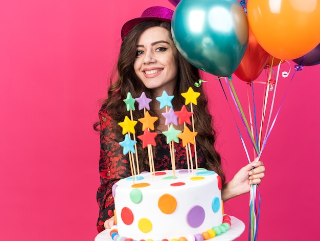 Smiling young party girl wearing party hat holding balloons and stretching out cake with stars towards camera looking at camera isolated on pink wall with copy space