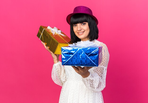 Smiling young party girl wearing party hat holding gift packages  isolated on pink wall with copy space