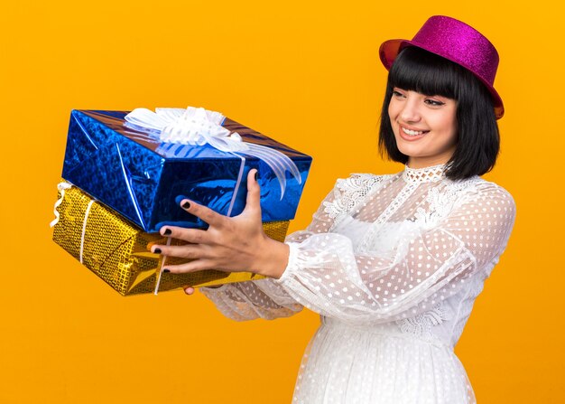 Smiling young party girl wearing party hat standing in profile view stretching out gift packages looking at them isolated on orange wall