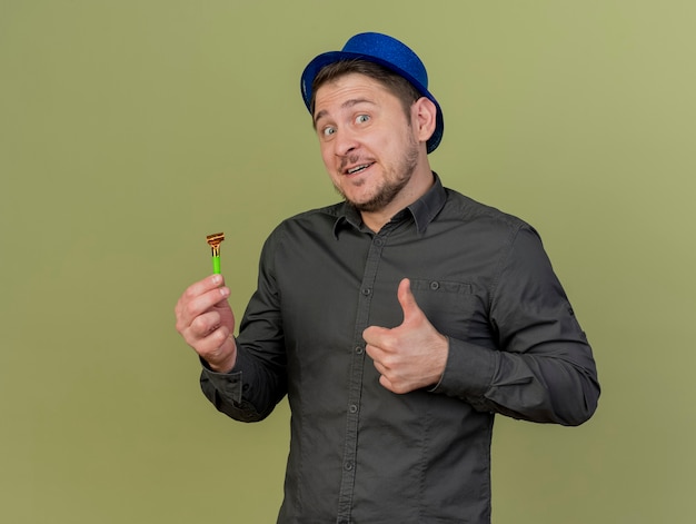 Smiling young party guy wearing black shirt and blue hat holding party blower showing thumb up isolated on olive green