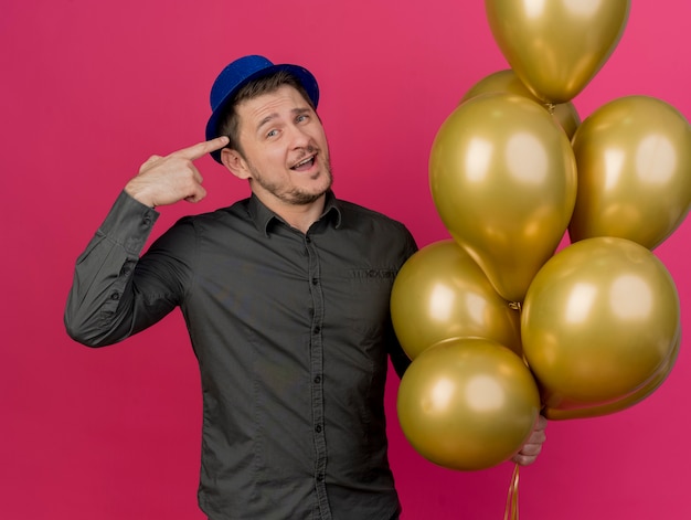 Smiling young party guy wearing blue hat holding balloons and points at himself isolated on pink