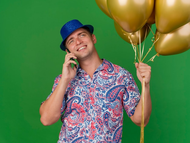 풍선을 들고 파란색 모자를 쓰고 웃는 젊은 파티 남자와 녹색에 고립 된 전화에 말한다