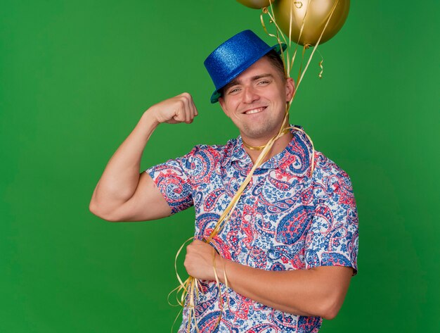 Smiling young party guy wearing blue hat holding balloons tied around neck isolated on green background