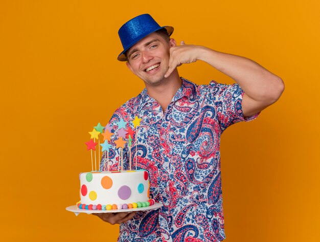 Smiling young party guy wearing blue hat holding cake and showing phone call gesture isolated on orange