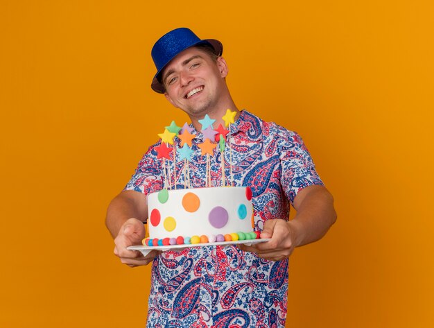 오렌지에 고립 된 케이크를 들고 파란색 모자를 쓰고 웃는 젊은 파티 남자