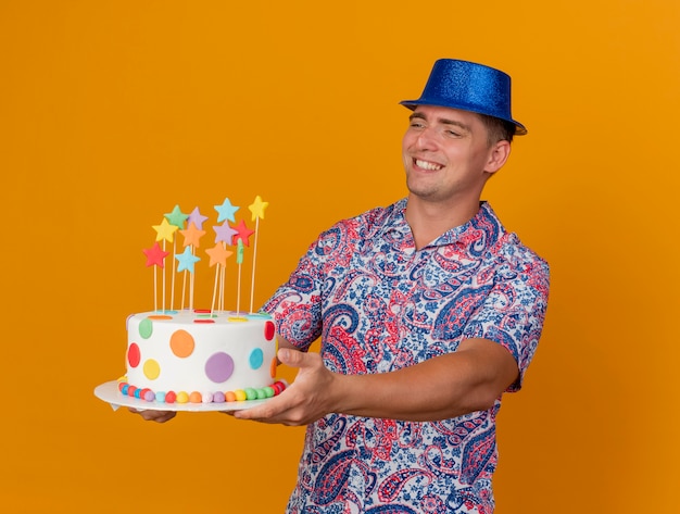 Smiling young party guy wearing blue hat holding out cake at side isolated on orange