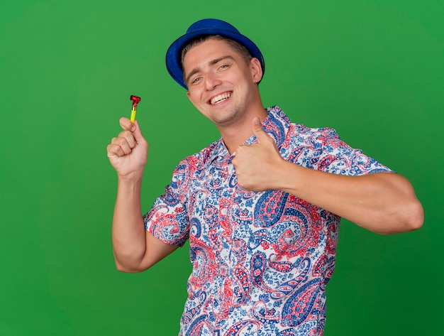 녹색에 고립 된 엄지 손가락을 보여주는 파티 송풍기를 들고 파란색 모자를 쓰고 웃는 젊은 파티 남자