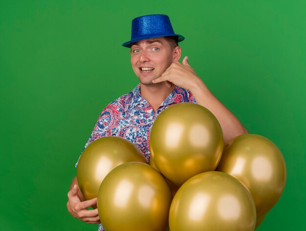 Smiling young party guy wearing blue hat standing behind balloons and showing phone call gesture isolated on green