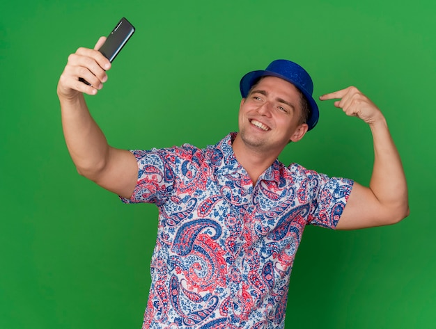 Foto gratuita il giovane ragazzo sorridente del partito che porta il cappello blu prende un selfie e indica se stesso isolato su priorità bassa verde