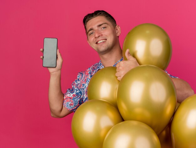핑크에 고립 된 엄지 손가락을 보여주는 풍선 가운데 서 화려한 셔츠를 입고 웃는 젊은 파티 남자