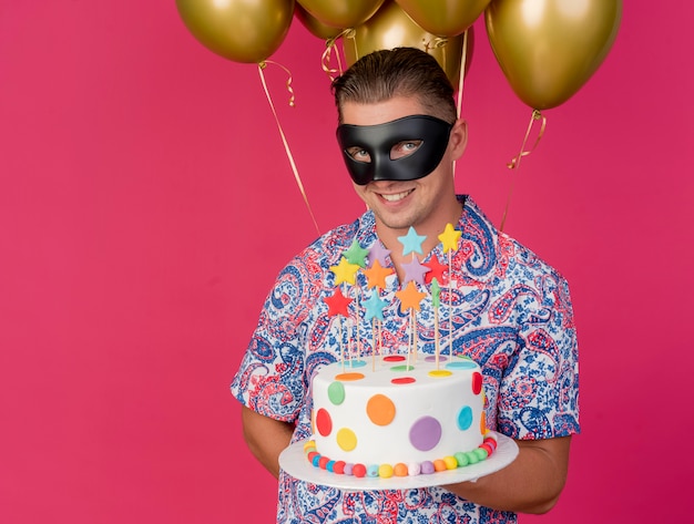 Ragazzo sorridente del partito giovane che indossa la maschera per gli occhi in maschera in piedi davanti a palloncini e tenendo la torta isolata sul rosa