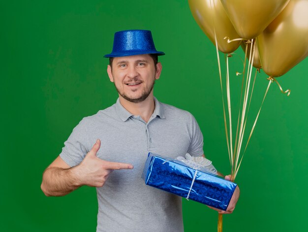 녹색에 고립 된 풍선 선물 상자에서 파티 모자 지주 및 포인트를 입고 웃는 젊은 파티 남자