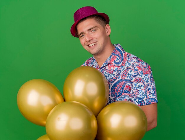 Smiling young party guy wearing pink hat holding balloons isolated on green