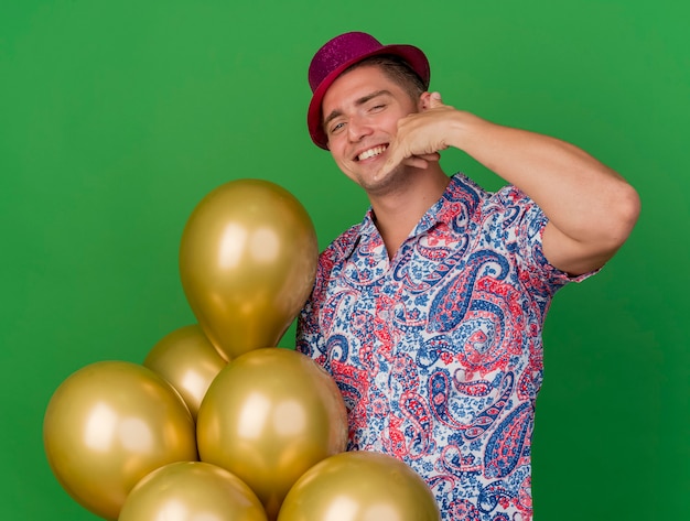 풍선을 들고 녹색에 고립 된 전화 제스처를 보여주는 분홍색 모자를 쓰고 웃는 젊은 파티 남자