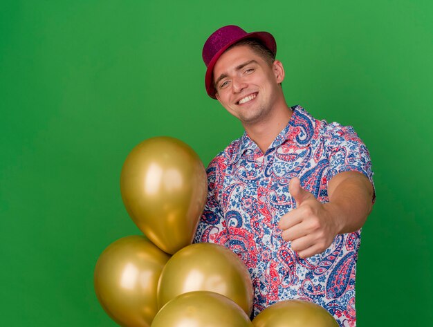 녹색에 고립 엄지 손가락을 보여주는 풍선을 들고 분홍색 모자를 쓰고 웃는 젊은 파티 남자