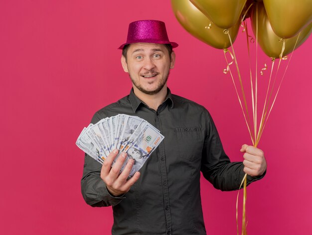 Smiling young party guy wearing pink hat holding balloons with cash isolated on pink