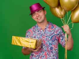 Бесплатное фото Улыбающийся молодой тусовщик в розовой шляпе держит воздушные шары с подарочной коробкой, изолированной на зеленом