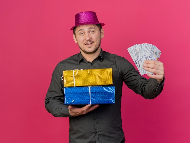 Ragazzo sorridente del partito giovane che indossa il cappello rosa che tiene i contenitori di regalo con contanti isolati sul colore rosa