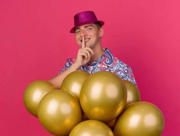 Smiling young party guy wearing pink hat standing behind balloons and showing silence gesture isolated on pink