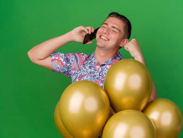 Smiling young party guy with closed eyes wearing colorful shirt standing behind balloons and speaks on phone showing yes gesture isolated on green