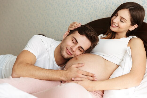 Улыбающаяся молодая беременная жена лежит в постели со своим мужем