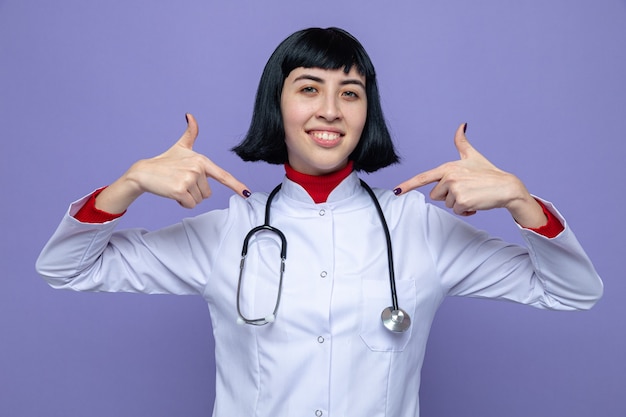 Smiling young pretty caucasian girl in doctor uniform with stethoscope pointing at herself 