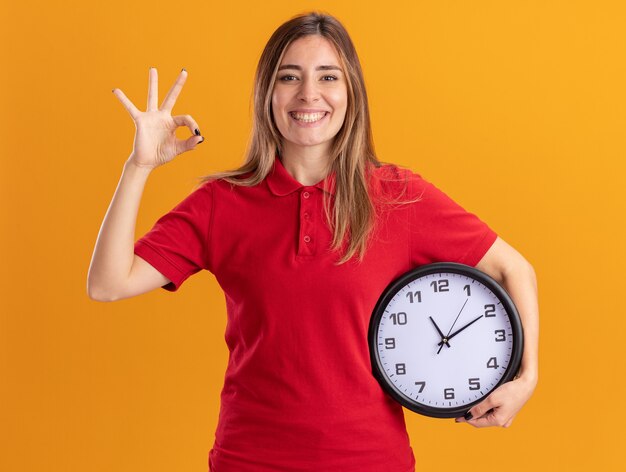 Smiling young pretty caucasian girl gestures ok hand sign and holds clock on orange