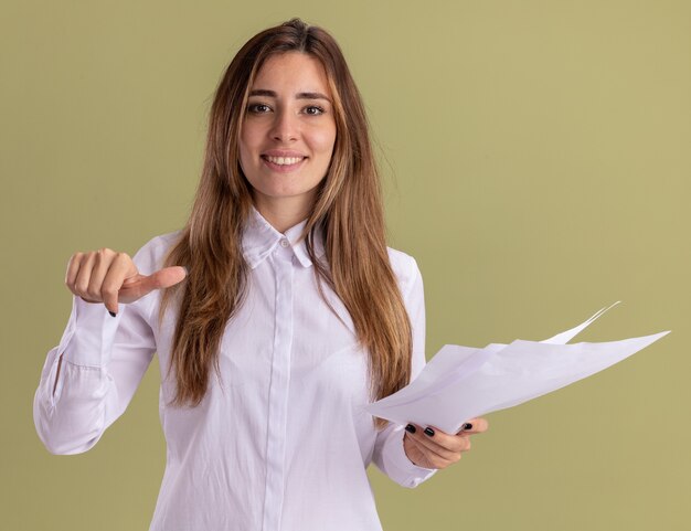 Smiling young pretty caucasian girl holds blank paper sheets and points at side isolated on olive green wall with copy space