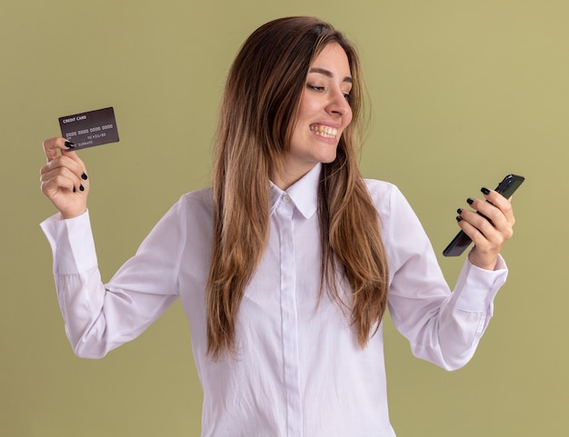 Smiling young pretty caucasian girl holds credit card and looks at phone isolated on olive green wall with copy space