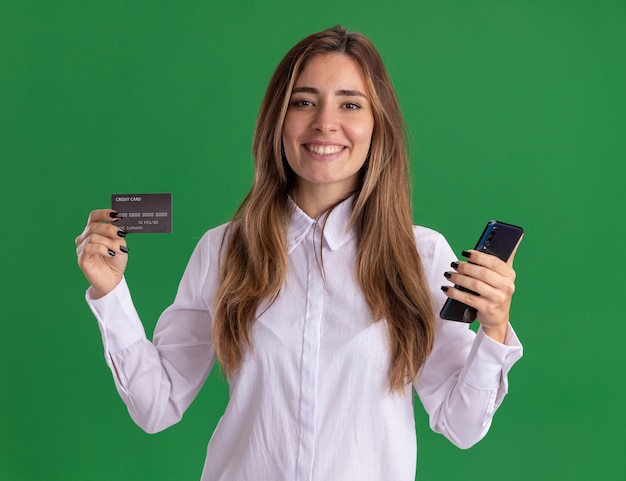 Free photo smiling young pretty caucasian girl holds credit card and phone on green