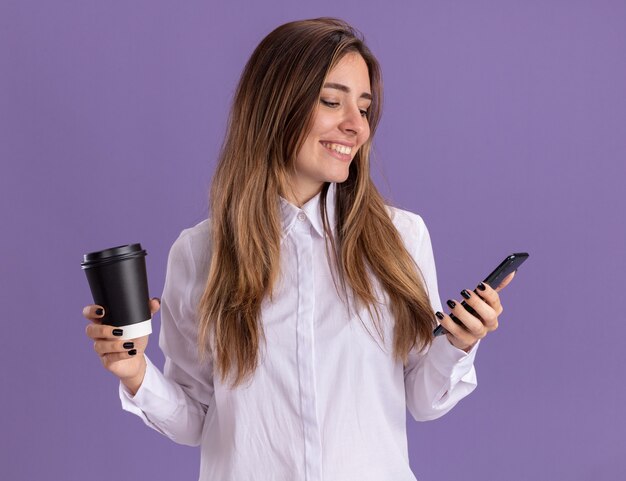 Smiling young pretty caucasian girl holds paper cup and looks at phone on purple 