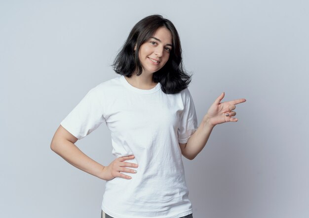 Smiling young pretty caucasian girl keeping hand on waist and pointing at side isolated on white background with copy space