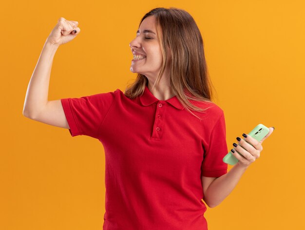 Smiling young pretty caucasian girl keeps fist and holds phone on orange